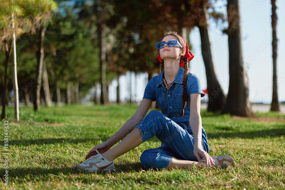 Fototapeta premium young woman sitting on grass outdoors wearing blue denim jumpsuit and sunglasses enjoying warm sunny day in green park environment