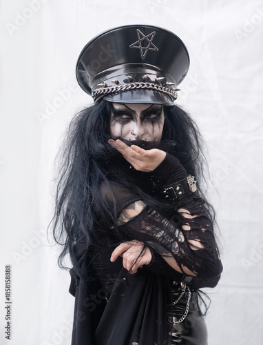 A gothic woman in black leather and shredded fabric poses with theatrical flair. Her face is painted dramatically, and a spiked military hat with pentagram crowns her bold, alternative look