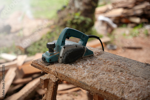 An electric wood planer resting on a rough wooden plank, covered in fresh sawdust, showing a paused woodworking process in an outdoor workshop environment.