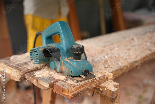An electric wood planer resting on a rough wooden plank, covered in fresh sawdust, showing a paused woodworking process in an outdoor workshop environment.
