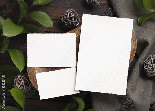 Blank cards on brown wooden tray near green leaves and grey fabric top view, boho mockup