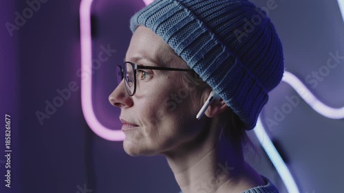Woman in profile wearing blue knit hat and glasses listening to music with earphones against dark purple-lit backdrop with neon-like lights
