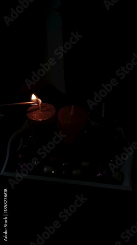 Close-up of a hand lighting three festive Christmas candles (gold, pink, and silver) with a match on a wooden table decorated with colorful ornaments. Warm holiday atmosphere in a dark room.