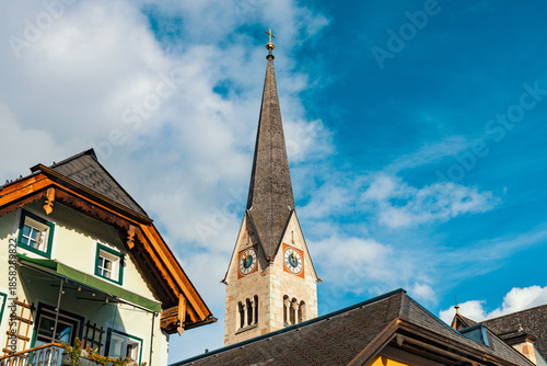 The slender, grey spire of the Hallstatt Lutheran Church rises elegantly against a bright blue sky, flanked by traditional alpine rooftops.