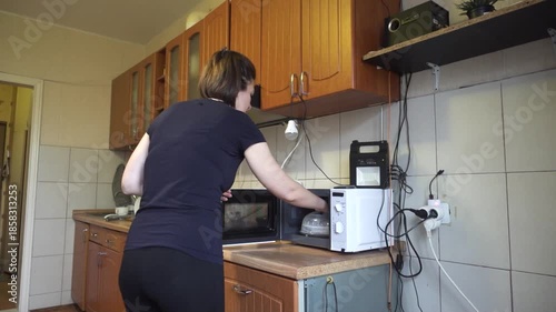 A woman opens a microwave in a cozy kitchen, illuminated by both natural light and a portable LED lamp. This image captures daily life and home cooking, hinting at energy solutions.