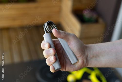 Close-up of a strong male hand squeezing a heavy-duty silver grip strengthener for armwrestling wrist training in a bright home gym. Background shows yellow bands, weight plates and fitness gear