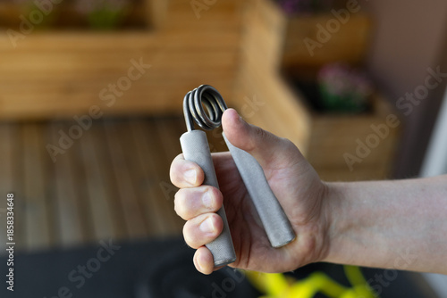 Close-up of a strong male hand squeezing a heavy-duty silver grip strengthener for armwrestling wrist training in a bright home gym. Background shows yellow bands, weight plates and fitness gear