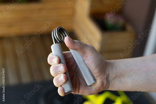 Close-up of a strong male hand squeezing a heavy-duty silver grip strengthener for armwrestling wrist training in a bright home gym. Background shows yellow bands, weight plates and fitness gear