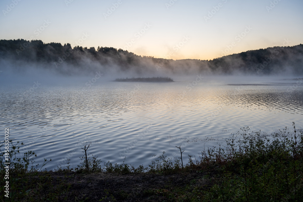 Fototapeta premium Golden morning mist rises from the calm waters of Lake Vlasina in Serbia, surrounding a small vegetated islet with a forested shoreline in the background.