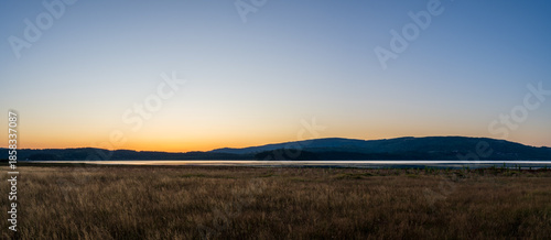 Minimalist dusk landscape featuring tall dry meadow grass, a reflective lake, and distant forested hills. 