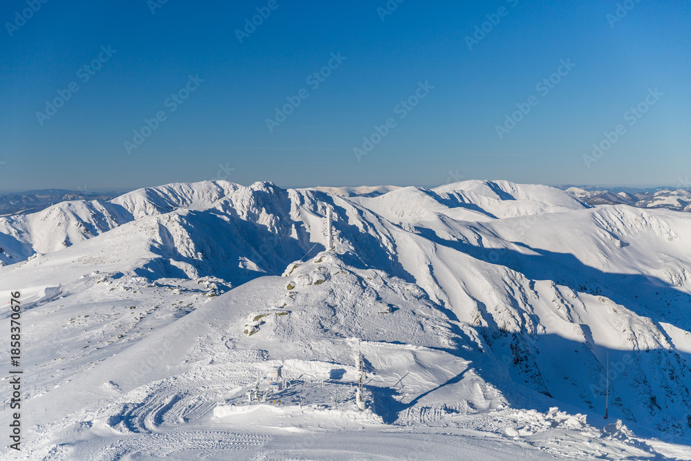 Fototapeta premium Wide panoramic view of snow-covered peaks in the Low Tatras National Park, Slovakia, featuring a bright sun star, frozen mountain ridges, and a distant view of the High Tatras under a clear winter sky
