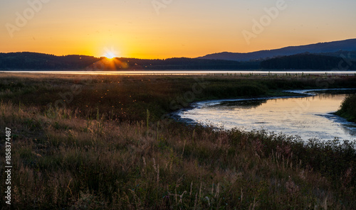 Golden sunset over a calm lake with wide open grassland in the foreground and dark rolling hills on the horizon. The clear sky fades from warm orange to soft blue.