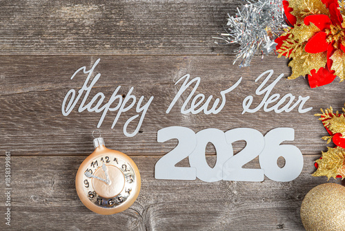 Happy New Year 2026 silver text on a weathered wooden background decorated with a clock bauble, Christmas ornaments, and vibrant red flowers.
