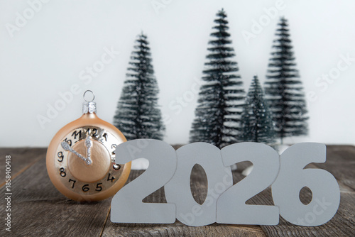 Silver 2026 numerals and a gold clock ornament set on a wooden surface with a blurred background of four snow-dusted miniature evergreen trees.
