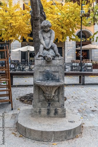 Font del Nen i la Tortuga historic fountain with boy and turtle statue at Independence Square. Autumn yellow leaves and cafe terrace in background on fall day. Girona, Spain - November 27, 2021