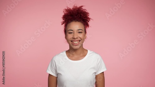 Close up smiling African American redhead curly woman in white t-shirt posing looks camera. Portrait positive female standing on isolated pink background with copy space. People emotions lifestyle