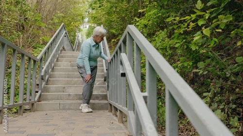 Elderly woman standing on outdoor stairs and holding her knee due to sudden pain