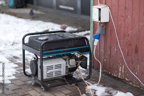 Portable gasoline generator connected to outdoor power socket near building during winter, emergency electricity supply concept.