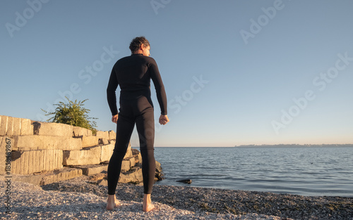 A man in a diving suit stands on the shore near the sea and looks into the distance, ready to enter the sea
