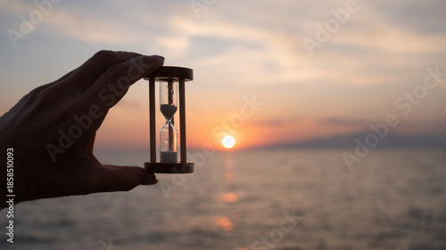 Hand holding hourglass against sea at sunset. Time management concept, personal control, reflection, future planning, new beginning, starting point, all over again and life transition