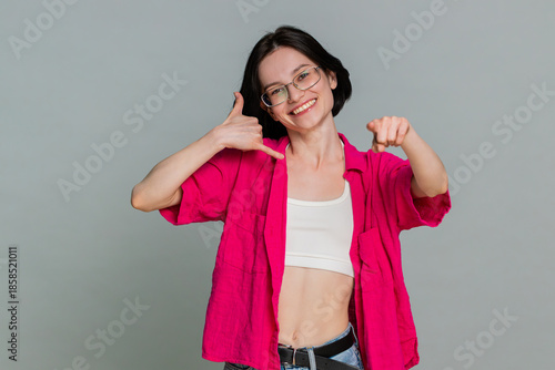 Young woman playfully lifts hand to ear with thumb and pinky finger extended, clearly making call me phone gesture. Caucasian girl expresses friendly communication request, isolated on gray background