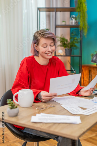 Young satisfied woman happily reviewing financial documents sitting at home table, smiling . Caucasian girl positively excited about good financial results, demonstrating successful money management.