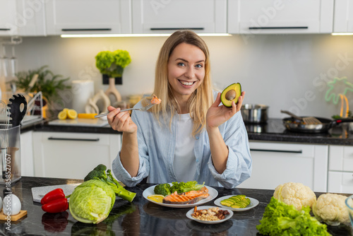Middle-aged woman eating trout fish with avocado nuts and vegetables for keto diet in kitchen at home feeling satisfied. Young girl choosing low carb meal for healthy fit nutrition and weight loss.