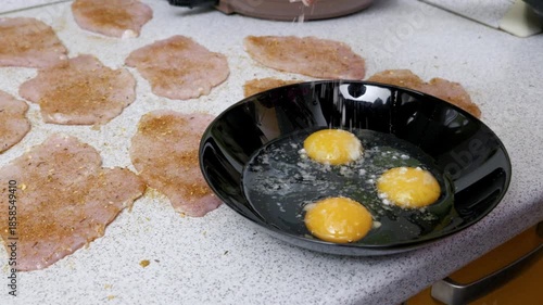 Salt is sprinkled into a black bowl with three raw eggs, prepared for breading seasoned meat cutlets. Close-up shot shows the food preparation process with natural kitchen lighting.