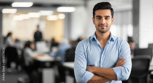 A confident young Indian businessman standing with arms crossed wearing a blue shirt in a bright, modern open-plan office background with copy space