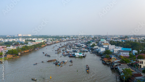 A wide aerial view captures the bustling Cai Rang floating market in Can Tho, Mekong Delta, southern Vietnam. Numerous boats gather along the river, surrounded by riverside houses and lush greenery