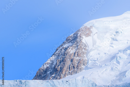 A dramatic rocky outcrop emerges from the snowy slopes and glacier of Mont Blanc du Tacul, highlighted by crisp winter sunlight and a clear blue sky.