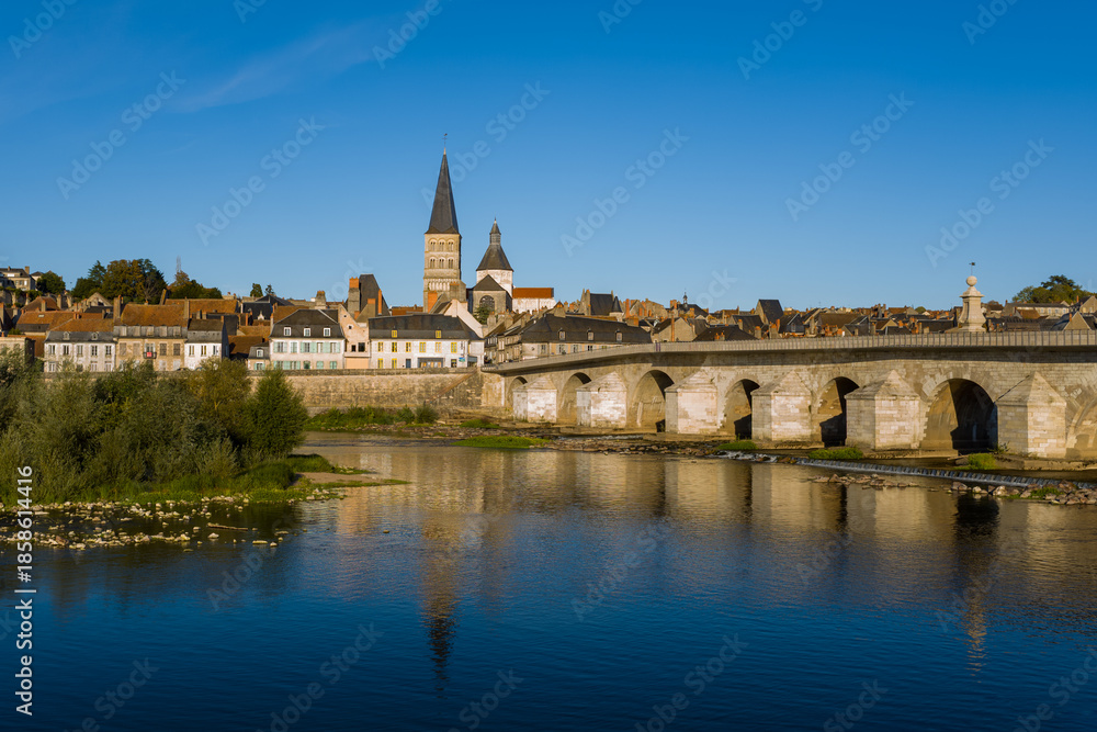 Obraz premium Under a vivid blue sky, the stone bridge and abbey spire of La Charite sur Loire rise above traditional riverside houses. Calm water reflects the town's medieval architecture and lush greenery