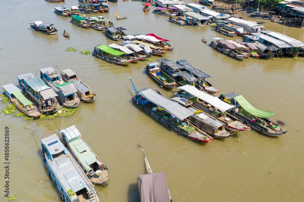 Obraz premium Aerial perspective reveals a lively cluster of covered market boats trading produce on the river at Cai Rang floating market in Can Tho, Mekong Delta, Vietnam. The scene features colorful tarps