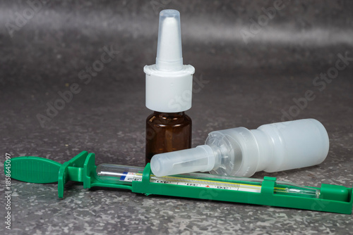The common medical supplies on a dark grey surface, featuring a brown glass nasal spray bottle, a white plastic dropper bottle, and a glass thermometer in a green protective case.