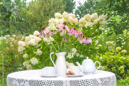 Summer breakfast in a blooming garden. Two white cups of tea, tea pot, bouquet of pink flowers on a table in a blooming garden