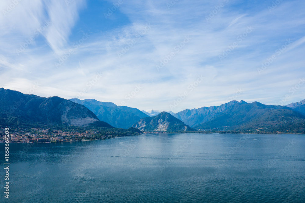 Obraz premium Wide view across the calm blue surface of Lake Maggiore, with dramatic mountain slopes and a lakeside town nestled at the base. Wispy clouds stretch across the sky, highlighting the expansive alpine
