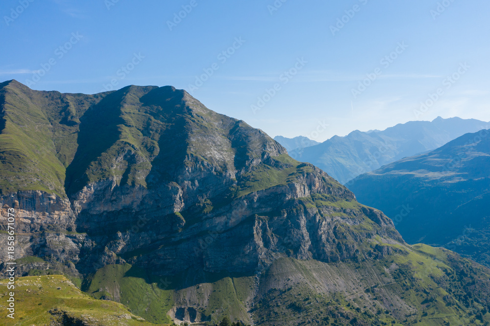Naklejka premium Majestic green and rocky cliffs rise sharply under a clear blue sky in the Pyrenees near Gavarnie Gedre. Layers of distant peaks fade into the horizon, creating a tranquil and expansive alpine scene.