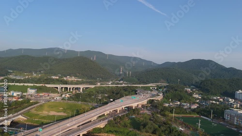 Aerial view of a modern highway system, including a prominent toll gate and multiple bridges, traversing a verdant valley flanked by tree-covered mountains with power lines.
