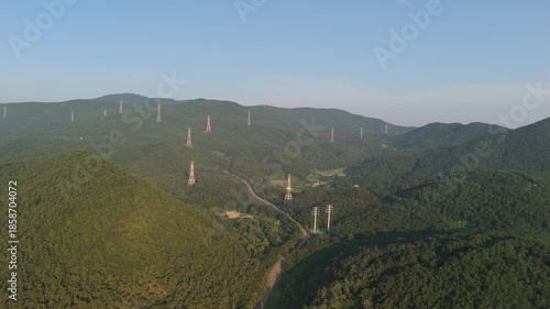 Aerial drone view: vast, densely forested mountains with numerous orange and white power transmission towers and lines. Winding road, buildings visible.