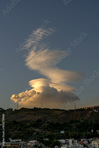 Dramatic lenticular cloud formation glowing in warm sunset light above a hillside town. Unique layered cloud structure rises into a clear blue sky, creating a powerful natural spectacle. 