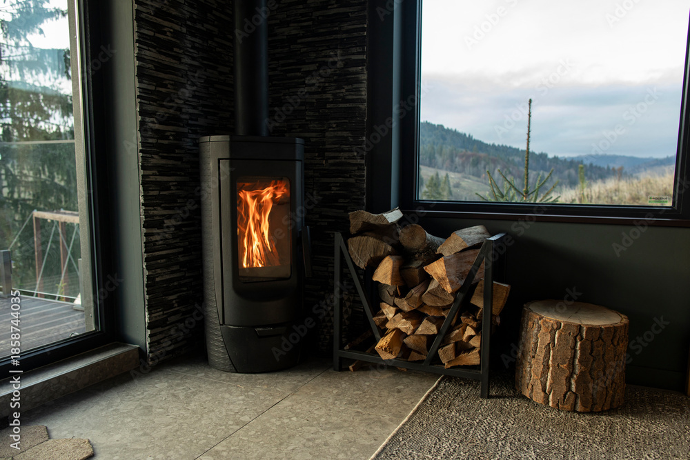 Fototapeta premium Modern wood stove burning brightly in cozy indoor setting near window, stacked logs nearby, view of mountains outside window creating warm atmosphere