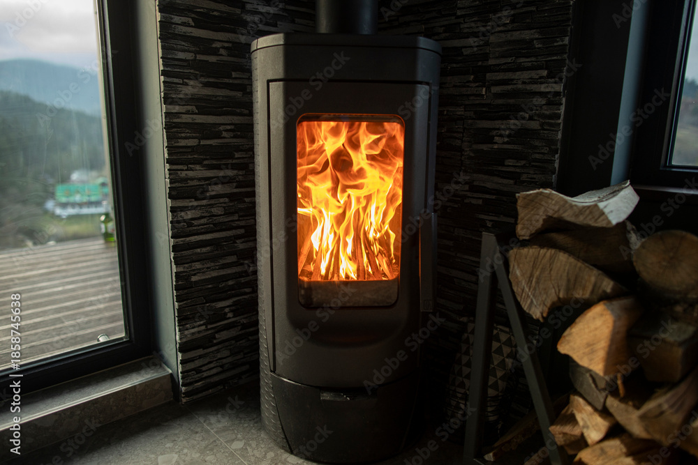 Fototapeta premium Wood stove burning brightly in the corner of cozy room, stacked logs nearby, large window with mountain view
