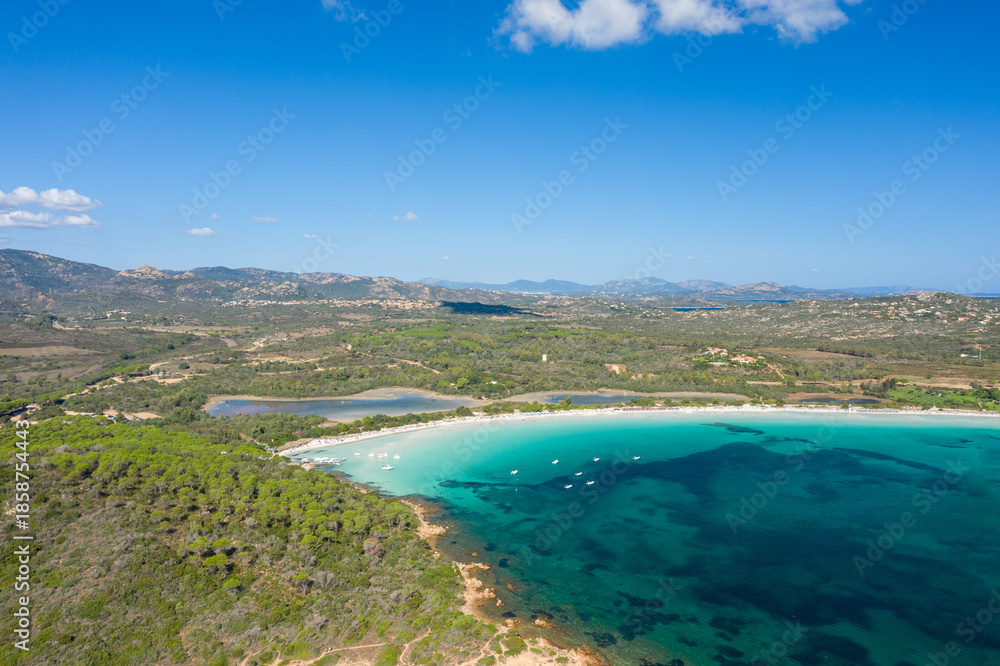 Fototapeta premium Aerial view of Cala Brandinchi in Sardinia, featuring a shallow turquoise lagoon, white sandy beach, and dense pine forest with rolling hills and distant mountains under a clear blue sky.