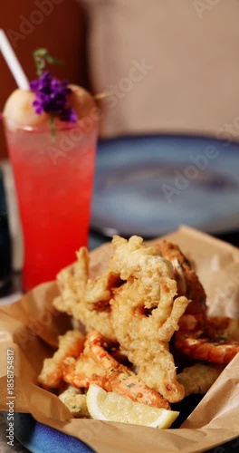 Crispy fried seafood, including shrimp tempura, served with lemon wedge in a restaurant. Gourmet dining experience. Appetizing, fresh, delicious food. Refreshing red cocktail.