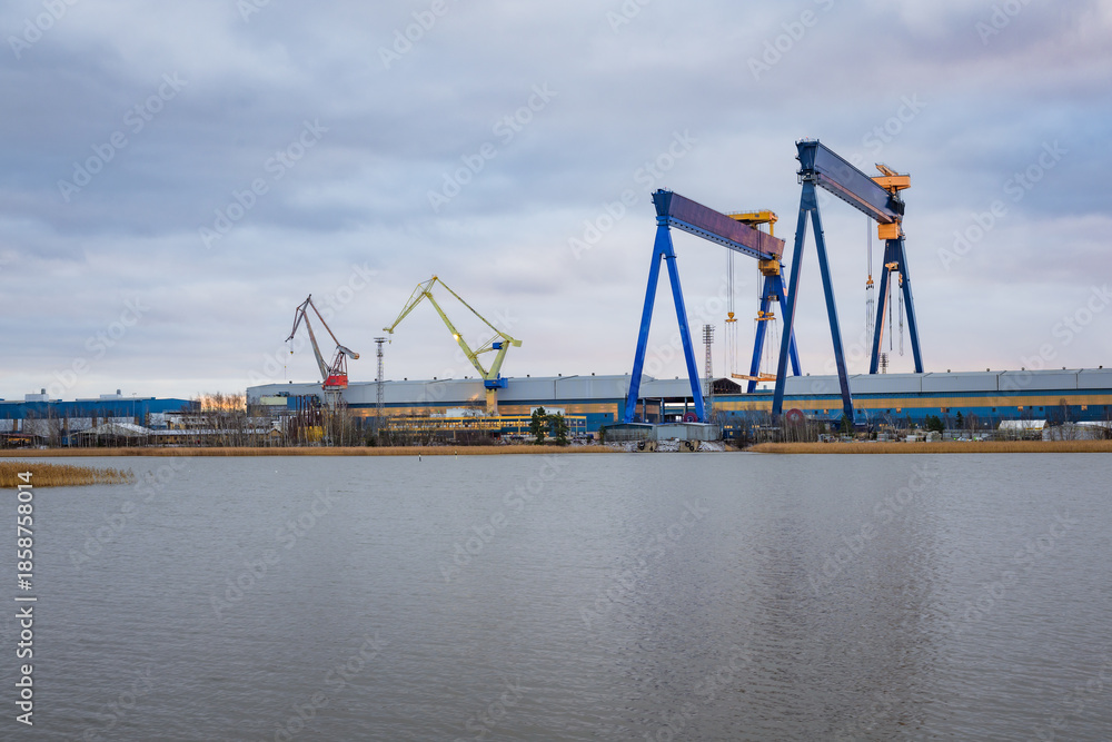 Obraz premium Photo of a large ship being constructed at a shipyard near the sea, captured from a distant vantage point. Shows cranes, scaffolding, massive vessel structure, and industrial maritime environment.