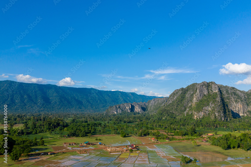 Fototapeta premium Aerial view of dramatic limestone cliffs rising above a mosaic of flooded rice fields and scattered rural houses, set against lush forest and distant mountains under a vivid blue sky. The landscape
