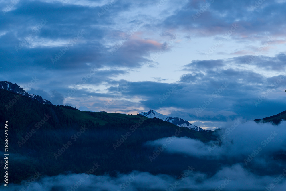 Naklejka premium Dramatic blue clouds drift above the snow-dusted summit of Mont Joly, with mist swirling over dark pine forests in the tranquil evening light.