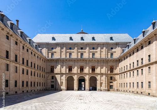 A sunlit symmetrical interior courtyard at El Escorial in Spain, featuring massive granite facades, arched walkways, and grey slate roofs under a clear blue sky