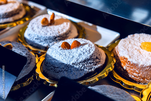 Trays cradle round Caprese cake dusted with powdered sugar, topped with almonds or candied fruit. Neat arrangement and rich texture glow under warm bakery lighting, evoking artisanal indulgence