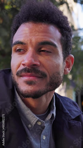 Vertical HD video. Portrait of young handsome bearded businessman standing outdoors in a city square, relaxing and looking around with a calm and thoughtful expression during a pleasant afternoon